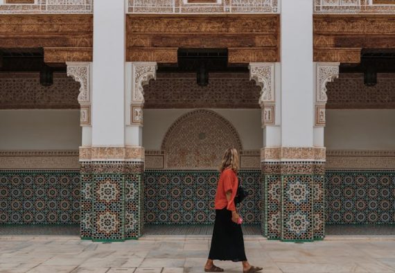 Woman walking past a decorated Moroccan facade and pool in Marrakesh, showcasing traditional mosaics.